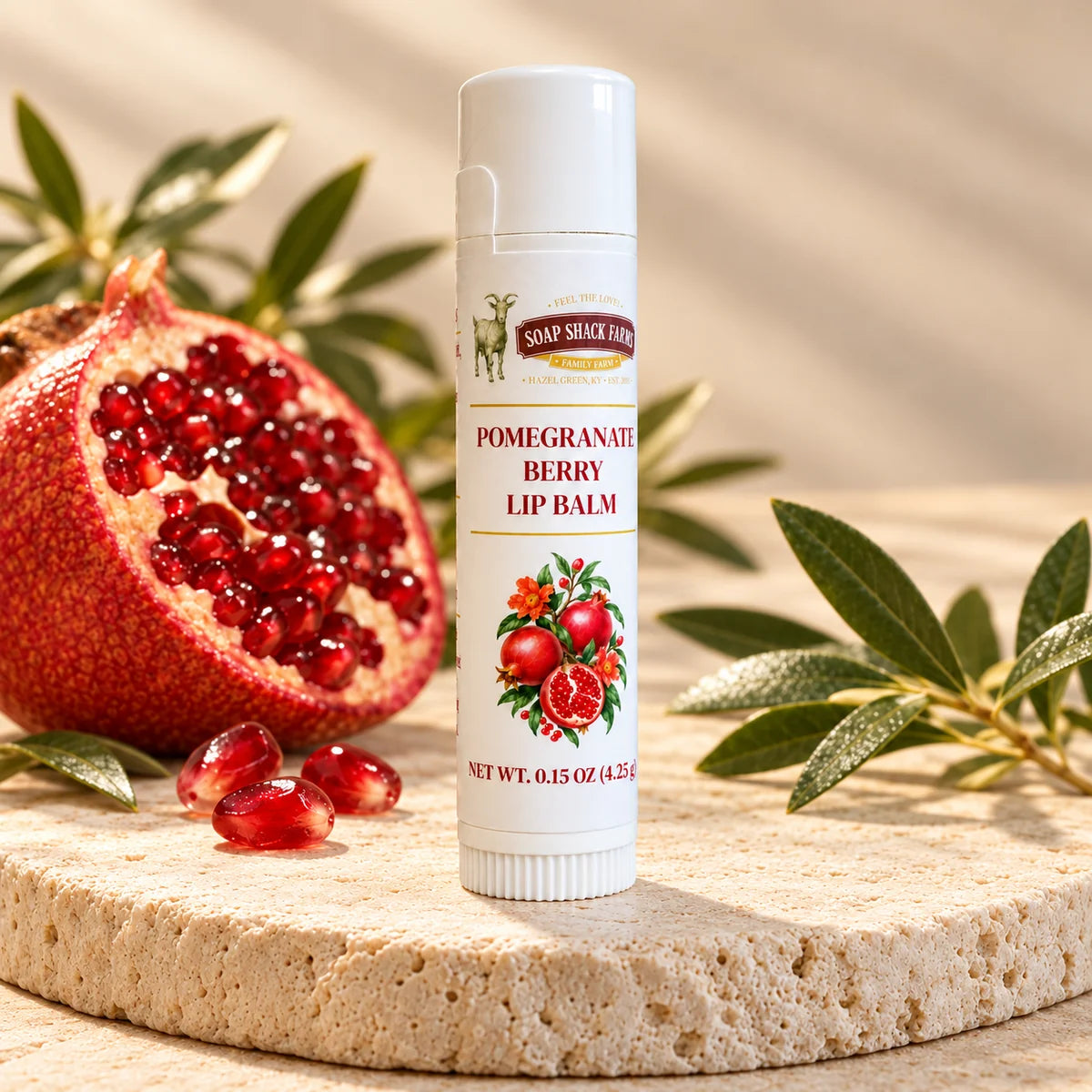 Soap Shack Farms Pomegranate Berry lip balm on a stone pedestal beside a cut pomegranate, seeds, and green leaves.