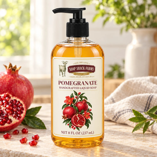 Pomegranate handcrafted liquid soap with fresh pomegranates, ruby seeds, greenery, and farmhouse linen in a sunlit setting.