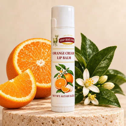 Soap Shack Farms Orange Cream lip balm on a stone pedestal beside orange slices, white citrus blossoms, and green leaves.