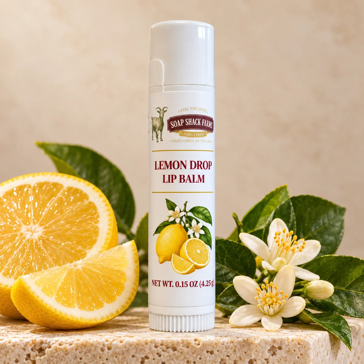 Soap Shack Farms Lemon Drop lip balm on a stone pedestal beside lemons, lemon slices, white citrus blossoms, and green leaves.