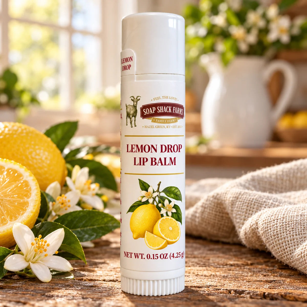 Soap Shack Farms Lemon Drop lip balm on a rustic wooden table beside lemons, citrus blossoms, and linen in a sunlit farmhouse kitchen.
