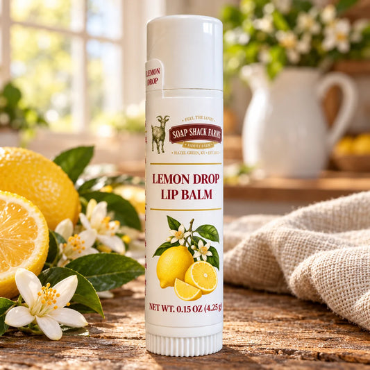 Soap Shack Farms Lemon Drop lip balm on a rustic wooden table beside lemons, citrus blossoms, and linen in a sunlit farmhouse kitchen.