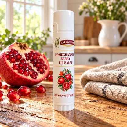Soap Shack Farms Pomegranate Berry lip balm on a stone pedestal beside a cut pomegranate, seeds, and green leaves.